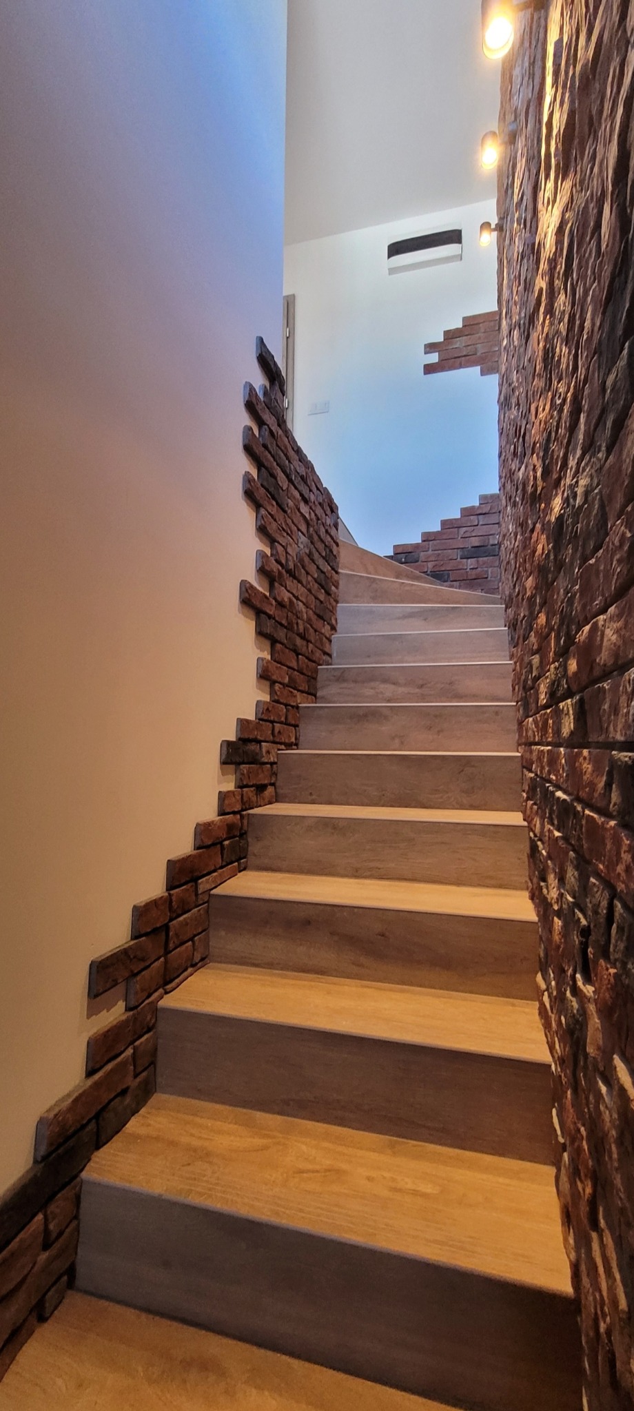 Schody z okładziną imitującą cegłę na ścianach, oświetlone punktowo. Widok z dołu, podkreślający detale wykończenia i fakturę materiałów. Nowoczesny design.