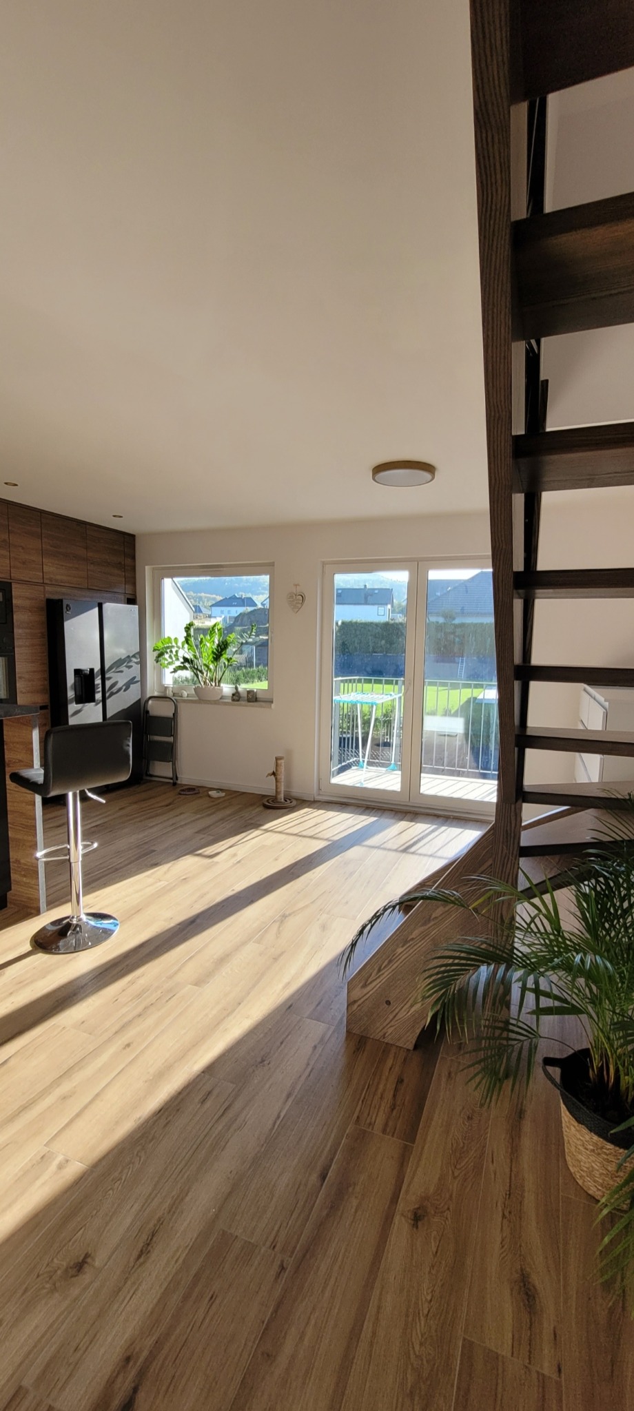 Jasne, przestronne wnętrze z drewnianą podłogą, schodami i dużymi oknami. Widoczne rośliny doniczkowe i krzesło barowe. Promienie słoneczne podkreślają ciepły charakter pomieszczenia.