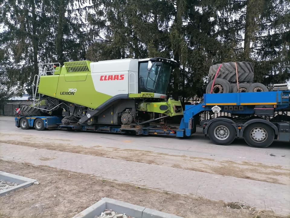 Kombajn zbożowy CLAAS Lexion 750 transportowany na niskopodwoziowej naczepie, dodatkowe opony zabezpieczone pasami na platformie. Widoczne drzewa w tle.