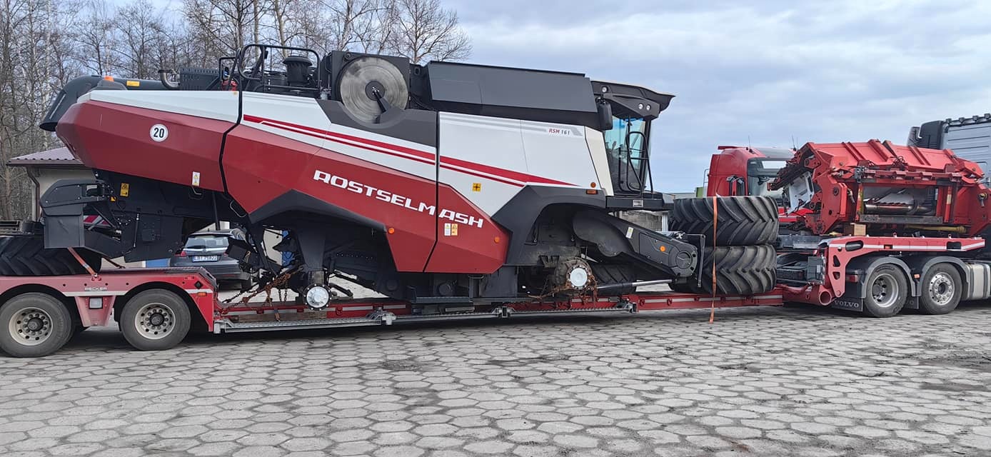 Czerwono-biały kombajn zbożowy ROSTSELMASH Rsm 161 przewożony na niskopodwoziowej platformie transportowej, z widocznymi elementami mocowania i oponami zapasowymi.
