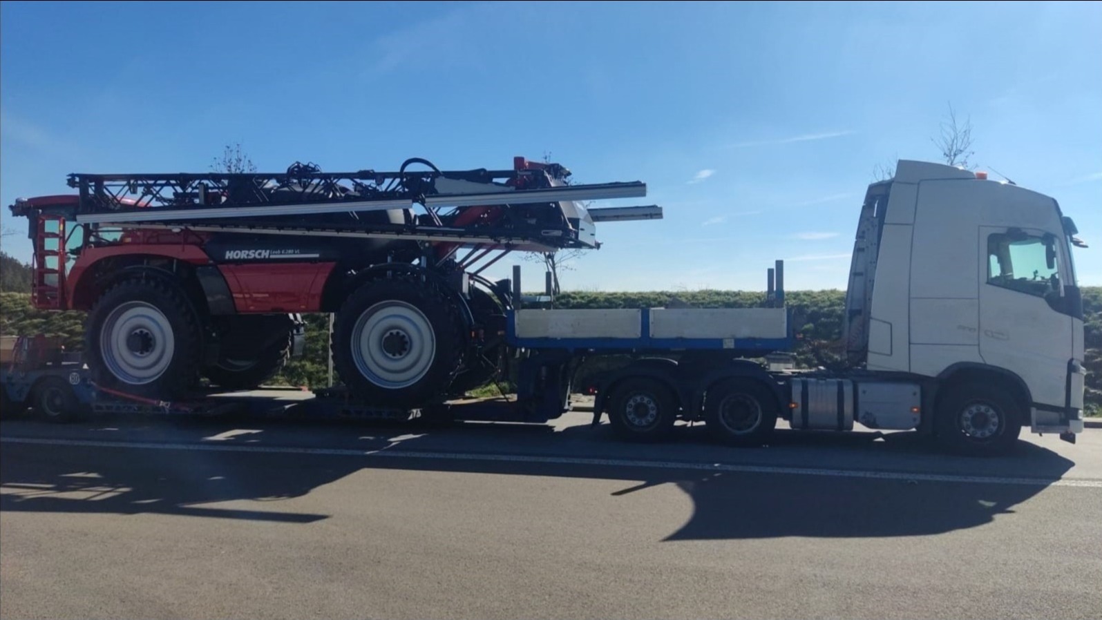 Czerwony opryskiwacz Horsch Leeb 6.280 VL na niskopodwoziowej naczepie ciągniętej przez białą ciężarówkę Volvo, transport drogowy w słoneczny dzień.