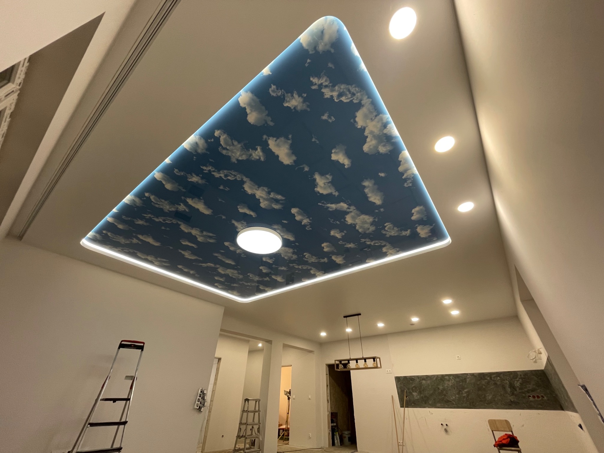 Nowoczesny pokój z trójkątnym, podświetlanym sufitem z motywem nieba i chmur, widoczne schody malarskie i przygotowania do dalszych prac wykończeniowych.