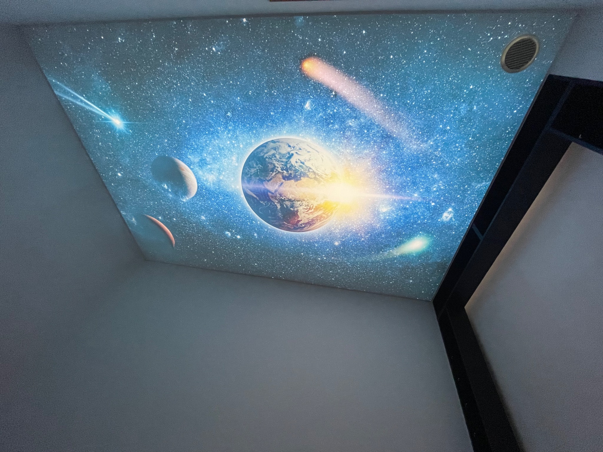 Pokój z sufitem przedstawiającym kosmos z planetami, gwiazdami i kometami, widoczna kratka wentylacyjna i fragment czarnej konstrukcji meblowej.