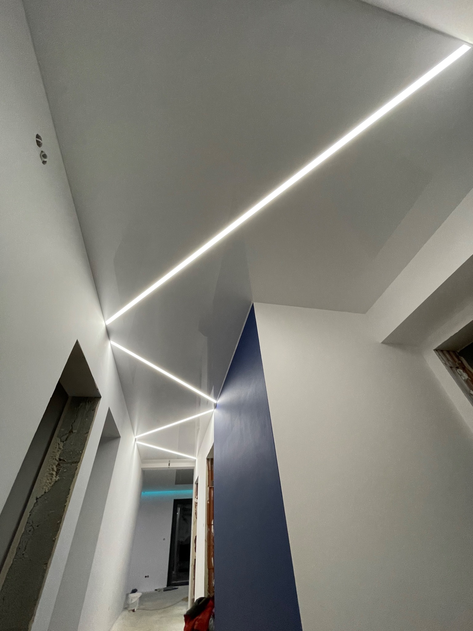 Biały sufit napinany z liniowym oświetleniem LED w kształcie zygzaka w korytarzu w trakcie remontu w Warszawie, widoczne niedokończone wnęki w ścianach.