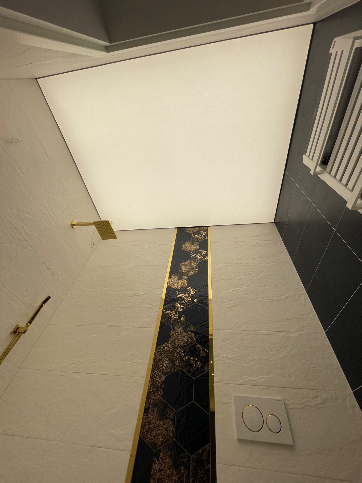 Nowoczesna łazienka z podświetlanym sufitem, złotą armaturą i dekoracyjnym pasem płytek z motywem kwiatowym w odcieniach czerni i złota.