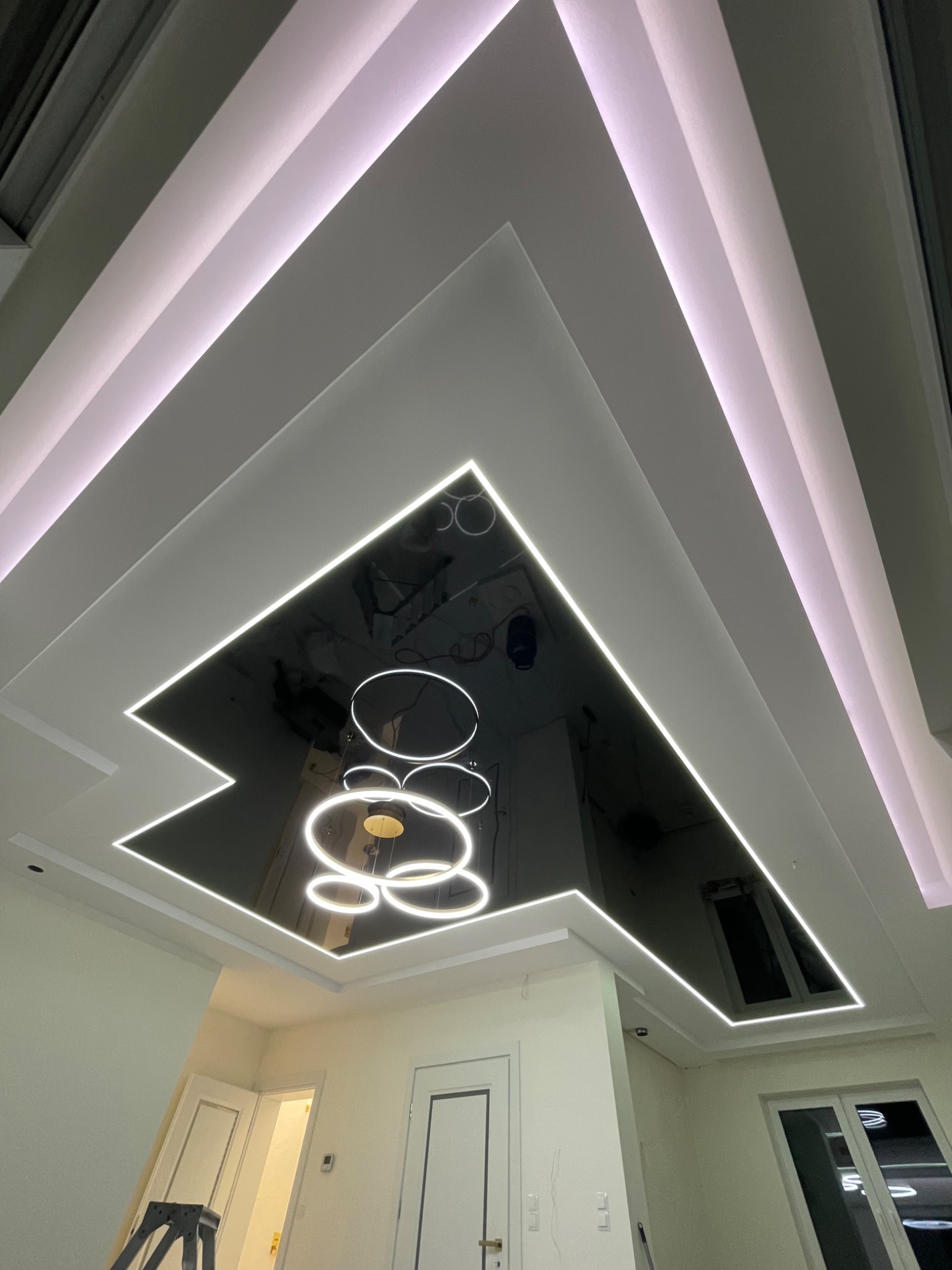 Wielopoziomowy sufit z czarną, lustrzaną powierzchnią w centralnej części, obramowany białymi listwami LED, nowoczesna lampa wisząca z okrągłych elementów, widoczny fragment drabiny malarskiej.
