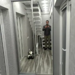 Wnętrze pokoju z szarą podłogą i ścianami, widoczna szafa przesuwna z lustrem, w której odbija się mężczyzna robiący zdjęcie telefonem, białe schody i okno balkonowe.