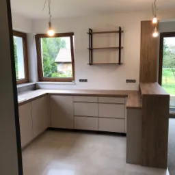 Jasna kuchnia na wymiar z drewnianym blatem i otwartymi półkami na ścianie, widok na ogród przez okno, surowy stan wykończenia z wiszącymi żarówkami.