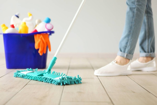 Osoba w jeansach i białych butach myje podłogę mopem z turkusową końcówką; w tle niebieskie wiadro ze środkami czystości i pomarańczowymi rękawicami.