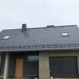 Dom w trakcie budowy z nowym, ciemnoszarym dachem z dachówki ceramicznej, widoczne okna dachowe i komin, elewacja ocieplona styropianem, częściowo otynkowana, brama garażowa w kolorze antracytowym.