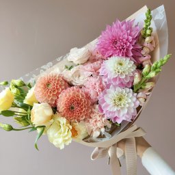 Design By Ann. Anna Supko - Pastelowy bukiet kwiatów z daliami, różami i goździkami, zawinięty w papier z motywem motyli, trzymany przez osobę w białym swetrze.