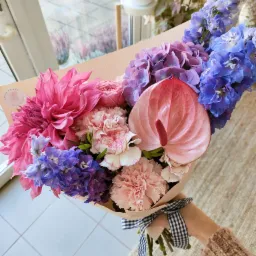 Bukiet różowych i fioletowych kwiatów, w tym dalii, anturium i goździków, owinięty papierem i przewiązany czarno-białą wstążką w kratkę, trzymany w dłoni ubranej w wełniany sweter.