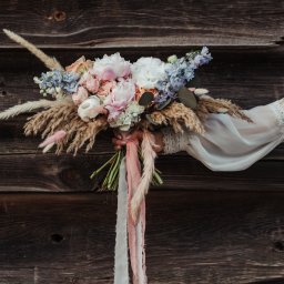 Design By Ann. Anna Supko - Bukiet ślubny z piwoniami, różami i trawami pampasowymi, trzymany przez osobę w koronkowej sukience na tle rustykalnej drewnianej ściany.