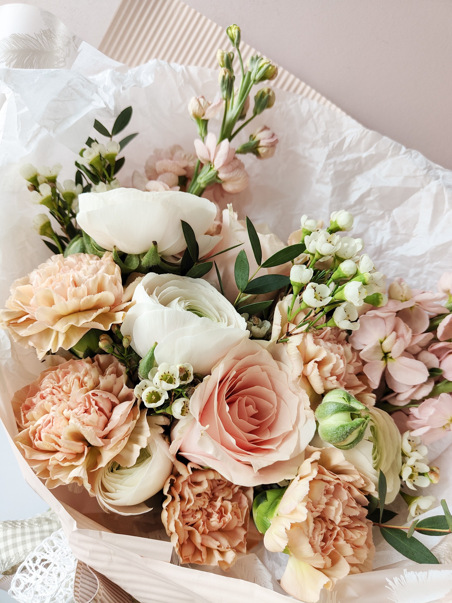 Delikatny bukiet z róż, goździków i ranunculus w pastelowych odcieniach różu i bieli, owinięty w biały papier i jasnoróżowy karton, z widocznymi zielonymi akcentami liści i pąków.