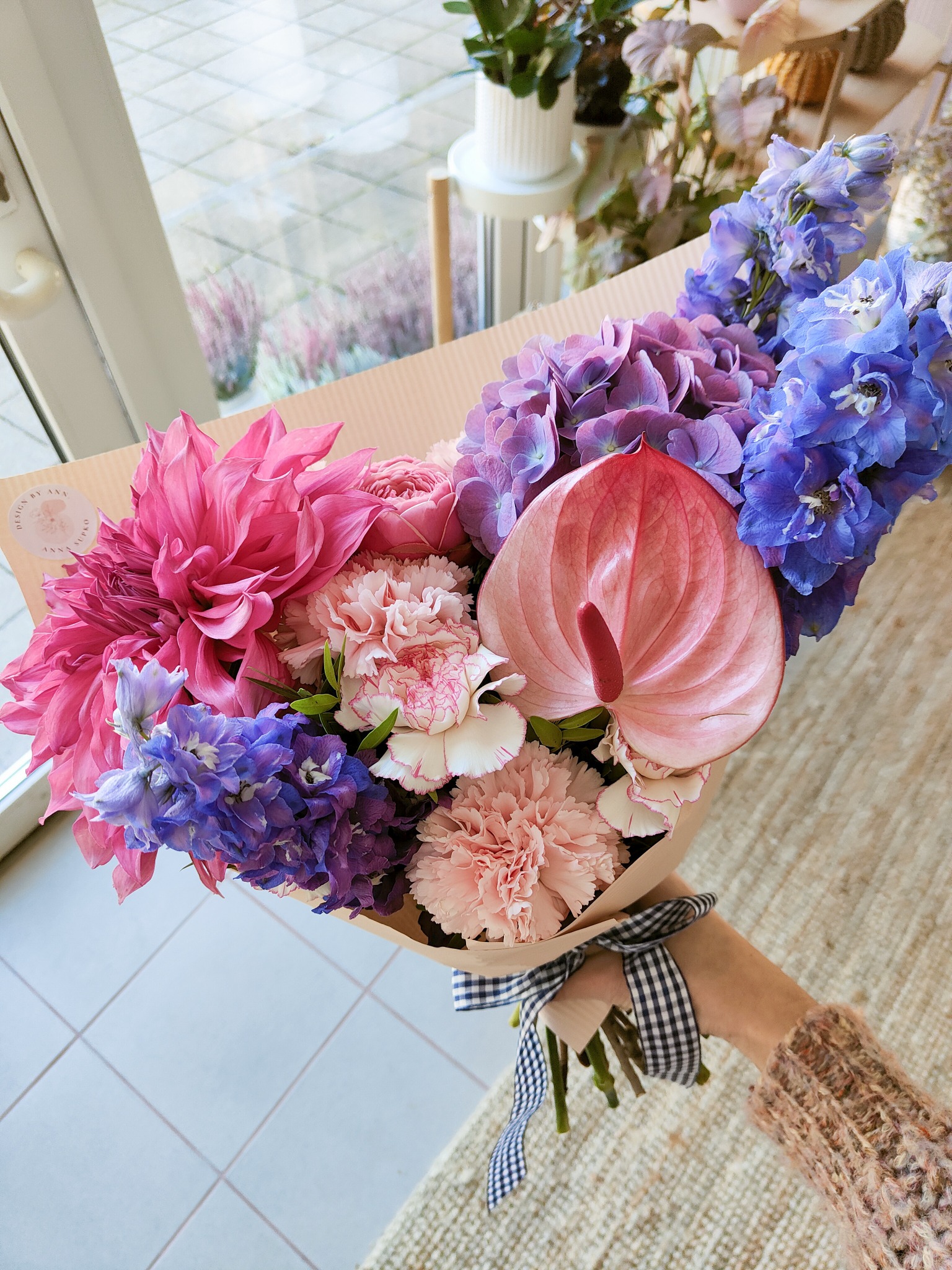 Bukiet różowych i fioletowych kwiatów, w tym dalii, anturium i goździków, owinięty papierem i przewiązany czarno-białą wstążką w kratkę, trzymany w dłoni ubranej w wełniany sweter.