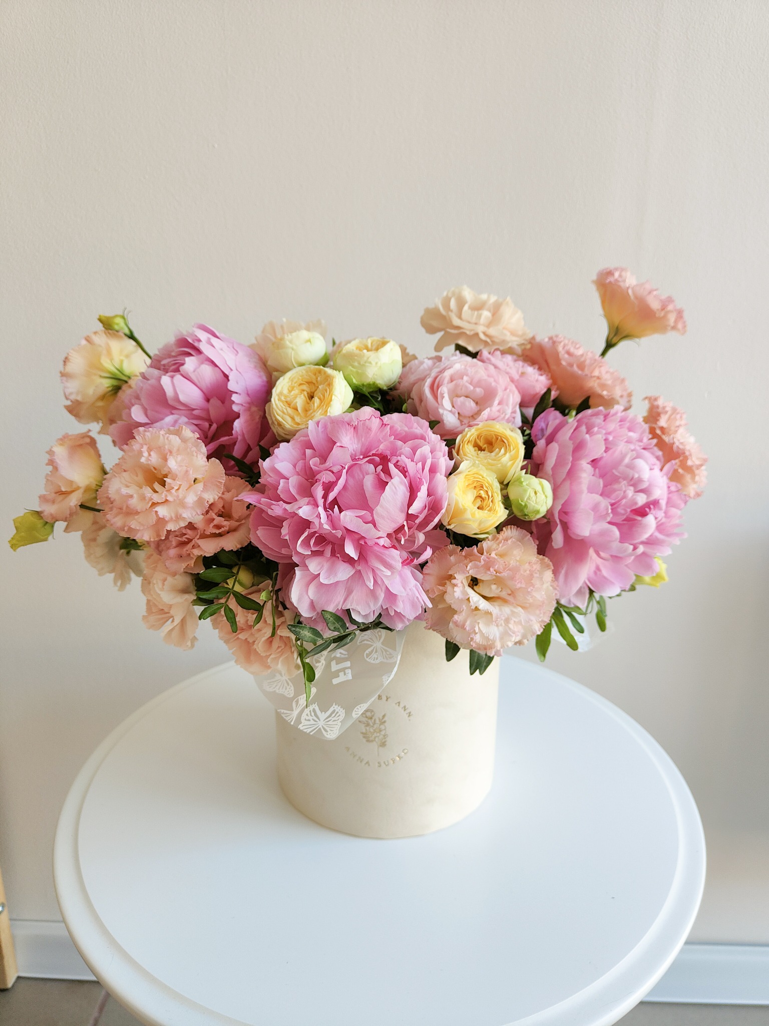 Kompozycja kwiatowa w okrągłym pudełku, zawierająca różowe piwonie, kremowe i żółte róże, całość umieszczona na białym stoliku na tle kremowej ściany.