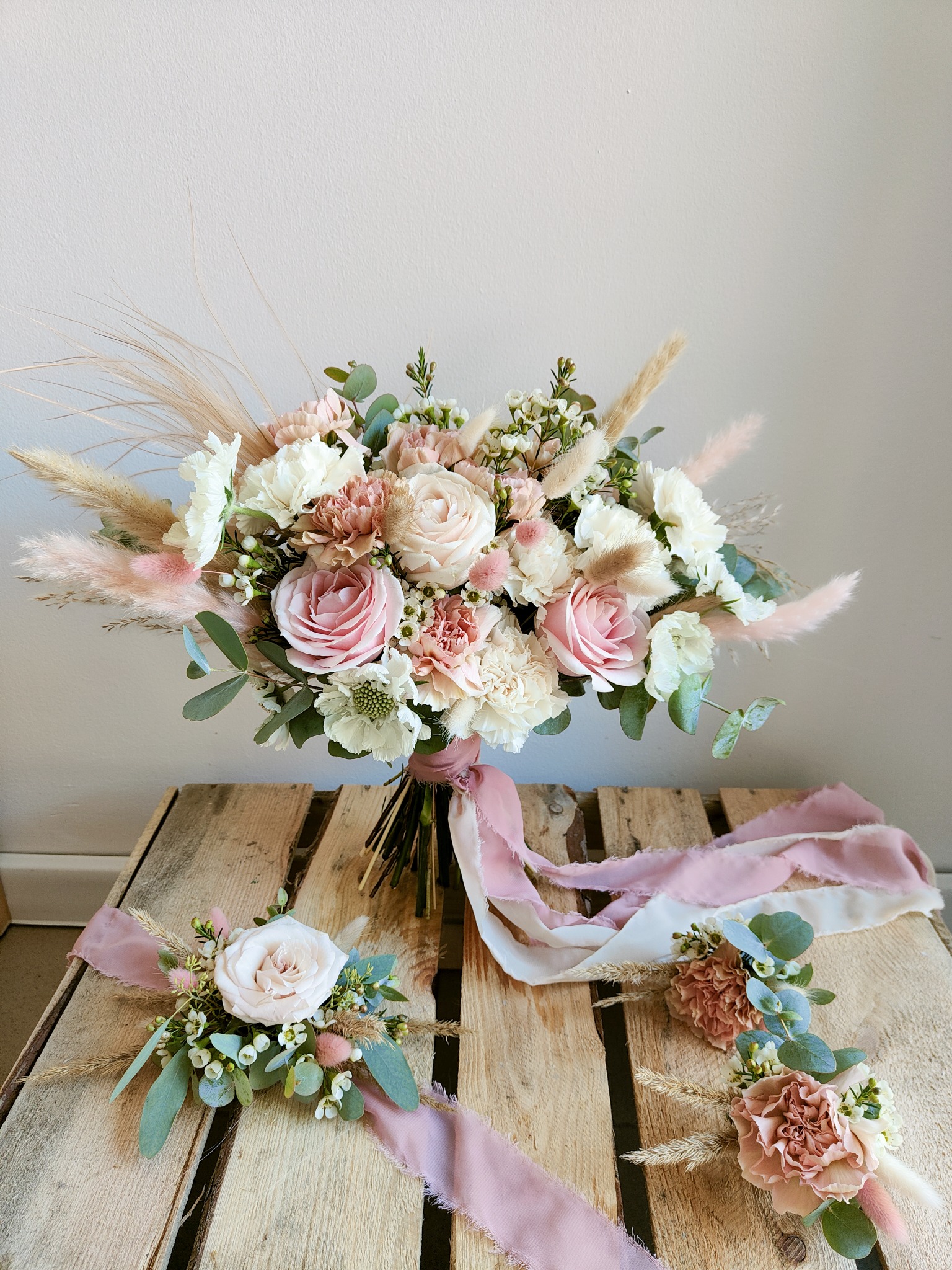 Delikatny bukiet ślubny z różowych i białych róż, goździków oraz traw pampasowych, uzupełniony o dwie butonierki i wstążki w pastelowych odcieniach różu, ułożony na drewnianej palecie.