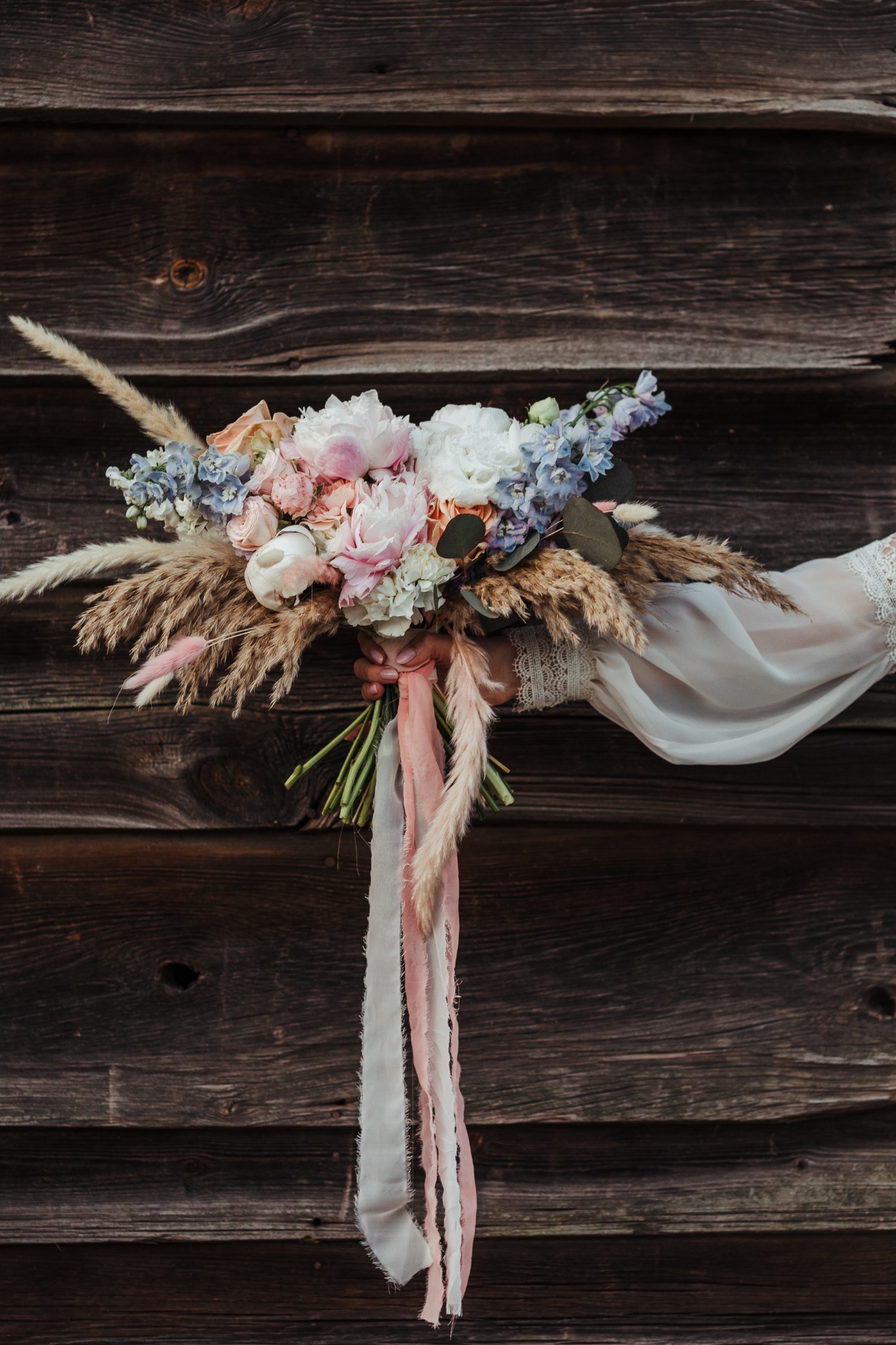 Bukiet ślubny z piwoniami, różami i trawami pampasowymi, trzymany przez osobę w koronkowej sukience na tle rustykalnej drewnianej ściany.