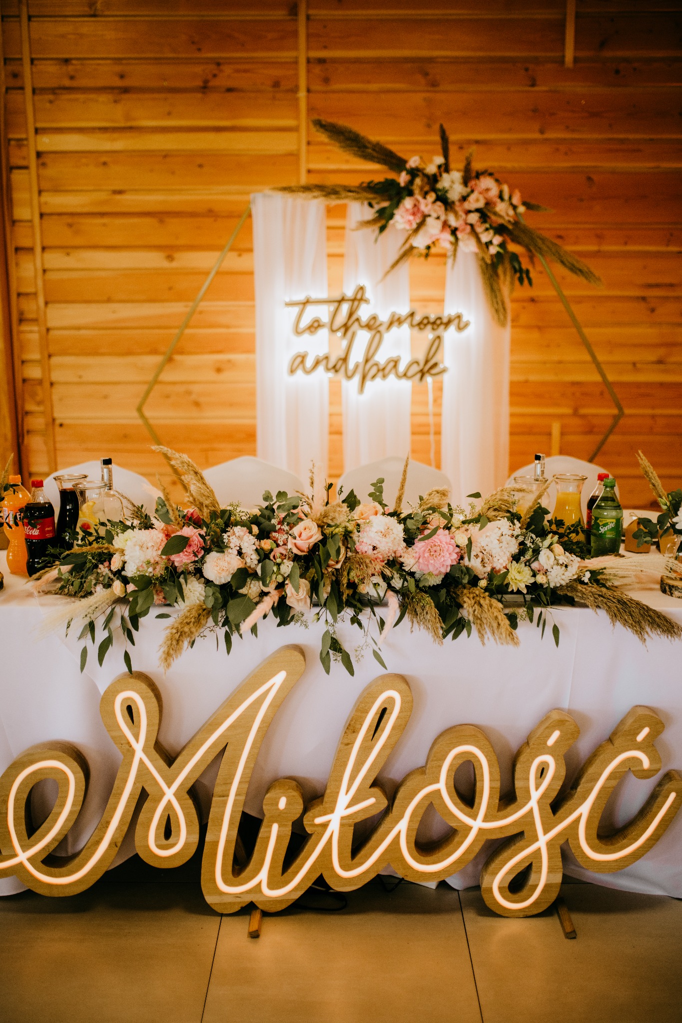 Dekoracja stołu weselnego z kwiatami, trawami pampasowymi i podświetlanym napisem 'Miłość' na pierwszym planie, w tle drewniana ściana i neon 'to the moon and back'.