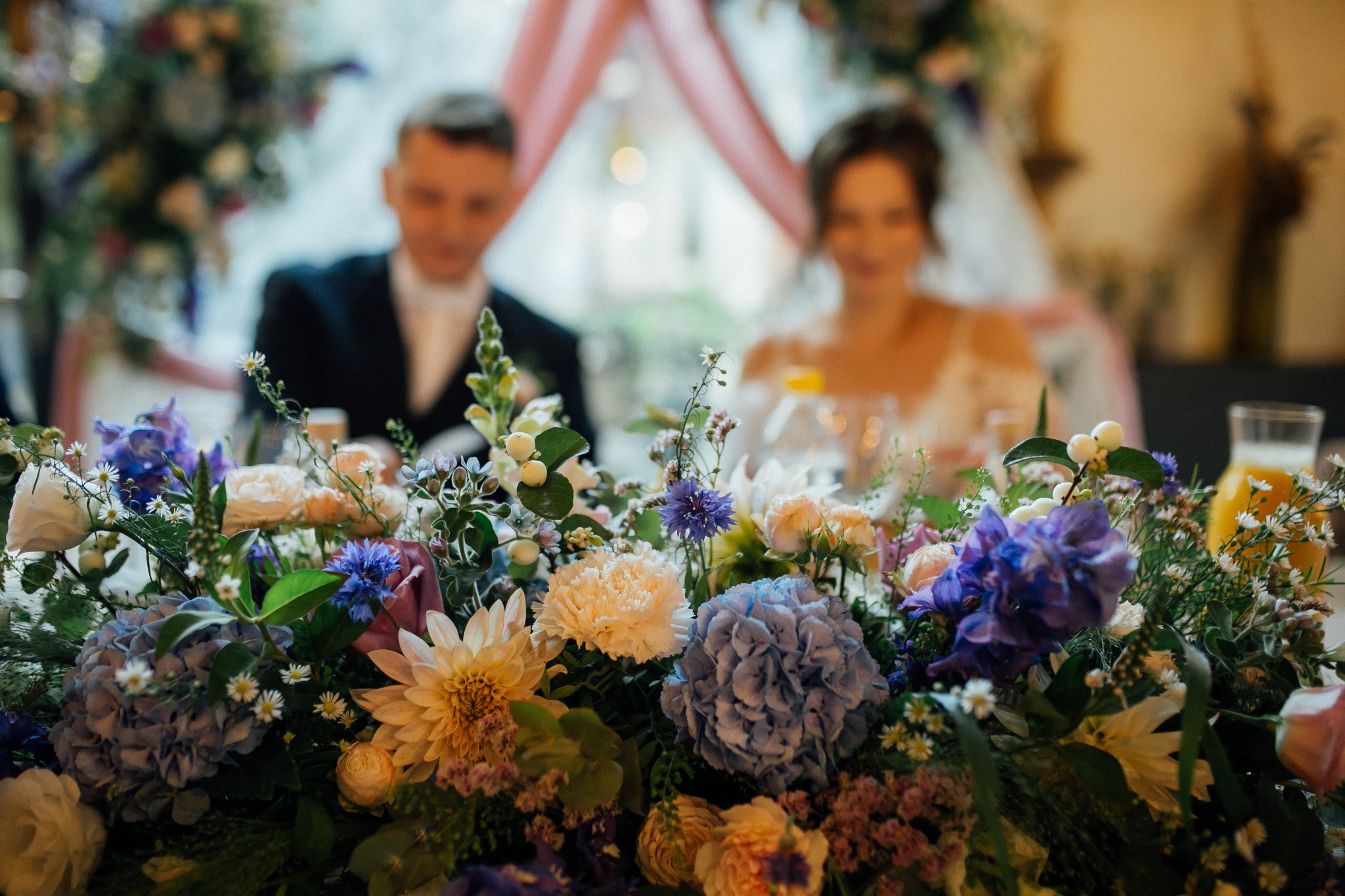 Dekoracja kwiatowa na stole weselnym z hortensjami, różami i chabrami w odcieniach bieli, różu i fioletu, w tle para młoda siedząca przy stole.