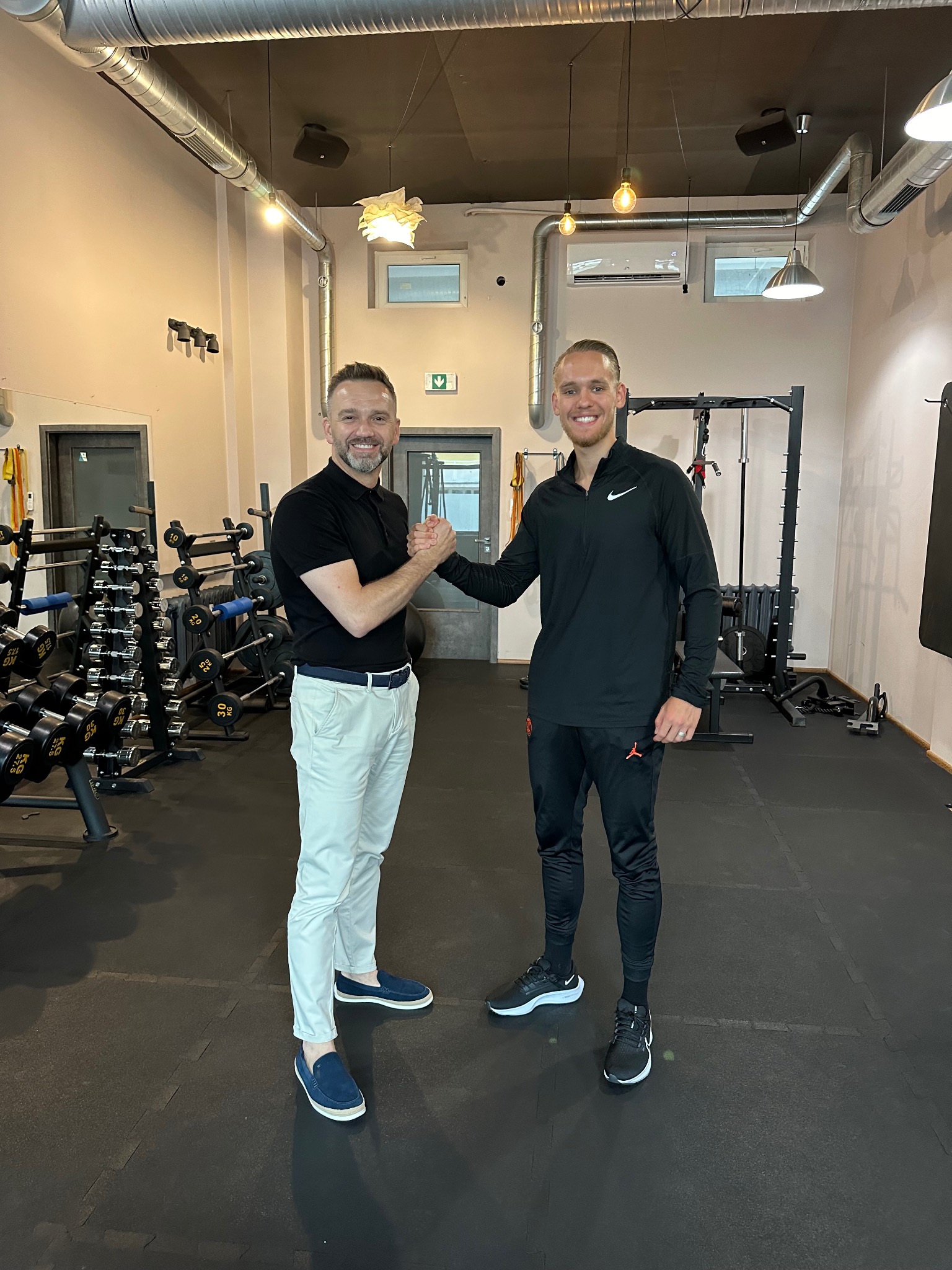 Dwóch mężczyzn w siłowni ściska sobie dłonie na tle sprzętu do ćwiczeń, widoczne hantle i maszyny.