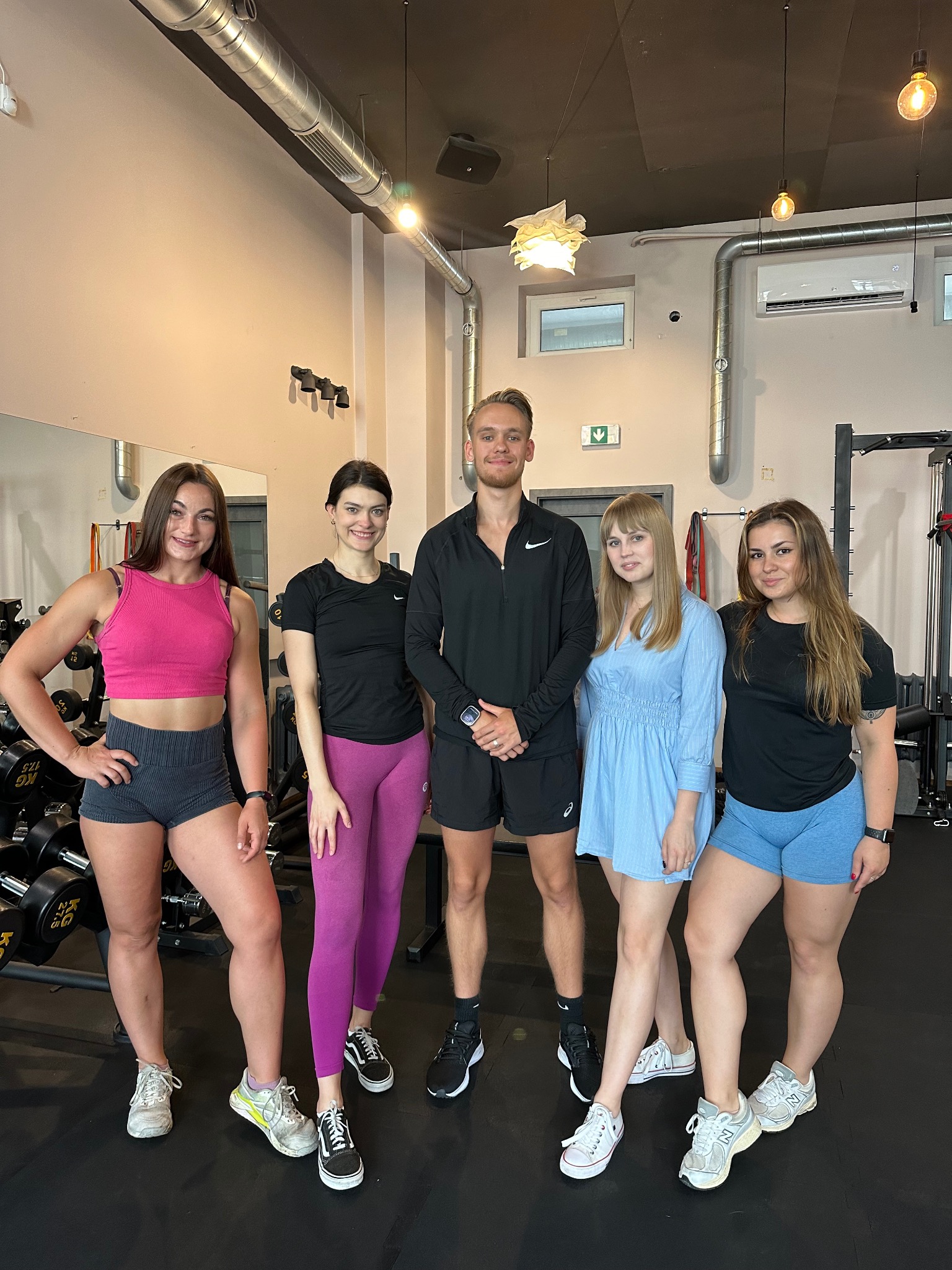 Grupa pięciu osób w siłowni, w tym mężczyzna w czarnej bluzie i spodenkach oraz cztery kobiety w strojach sportowych, pozujących na tle sprzętu do ćwiczeń.