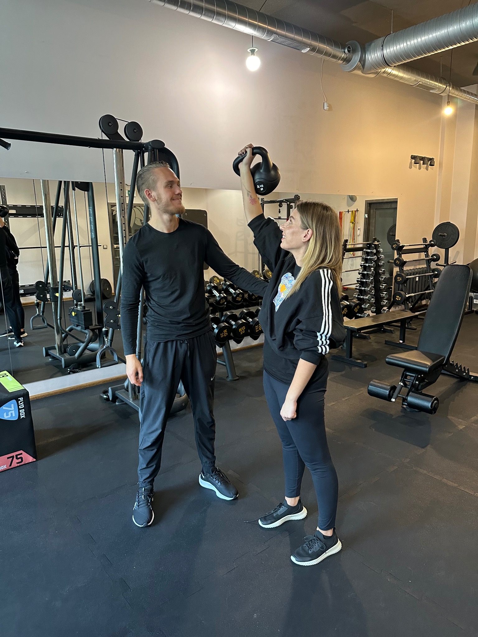 Kobieta podnosi kettlebell nad głowę z pomocą mężczyzny w siłowni, widoczne maszyny do ćwiczeń i ciężarki.