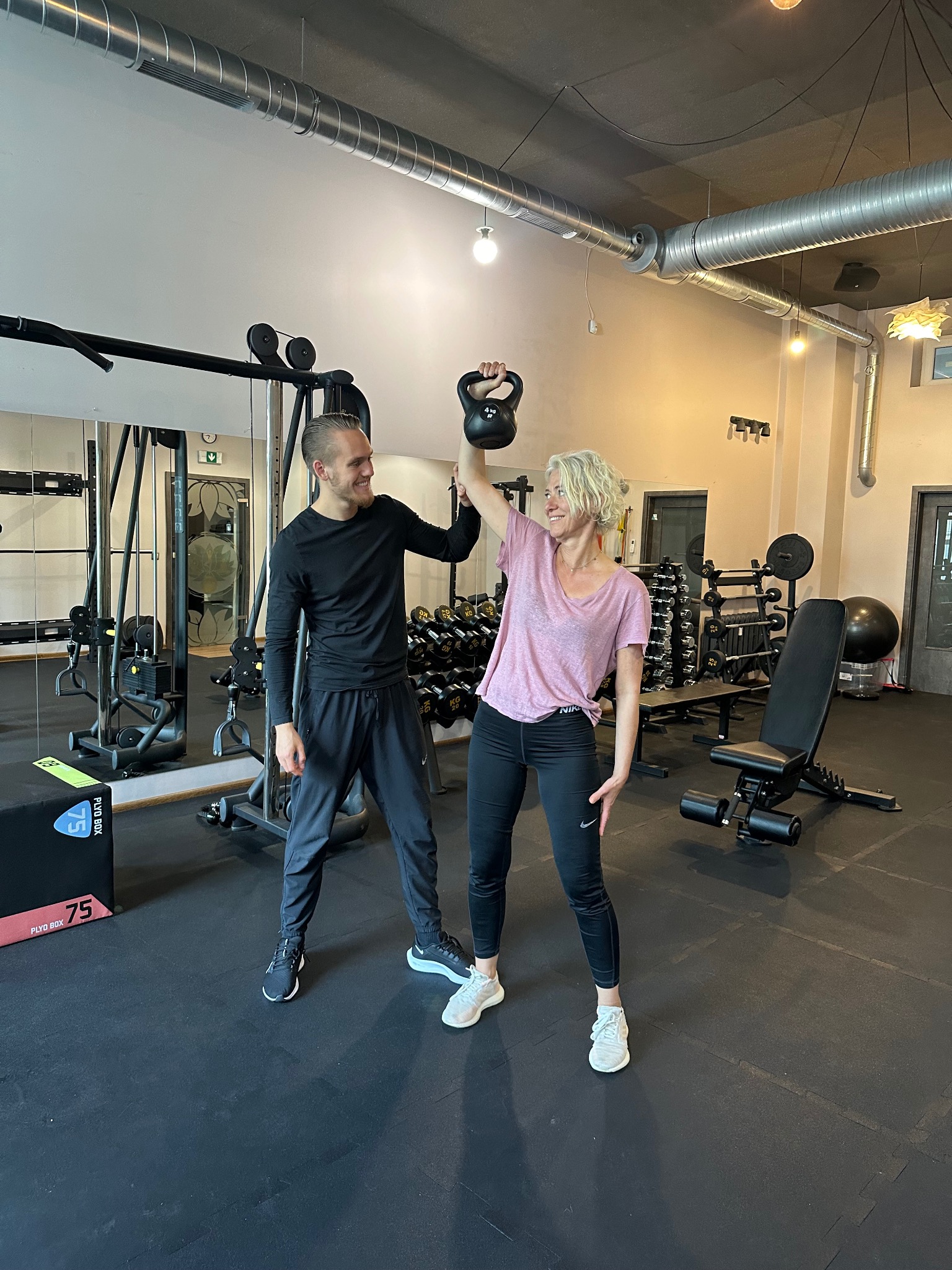 Kobieta z blond włosami podnosi kettlebell nad głowę, uśmiechając się do trenera w czarnym stroju sportowym w tle siłowni z różnymi sprzętami do ćwiczeń.