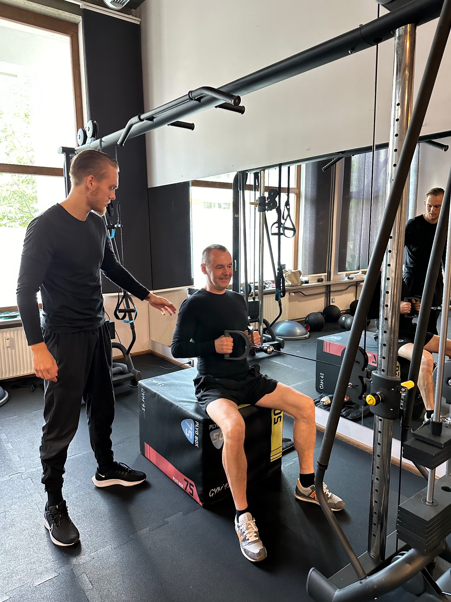 Mężczyzna ćwiczy na maszynie siłowej pod okiem instruktora w jasnej siłowni z lustrami i oknami, siedząc na czarno-czerwonym boksie plyometrycznym.