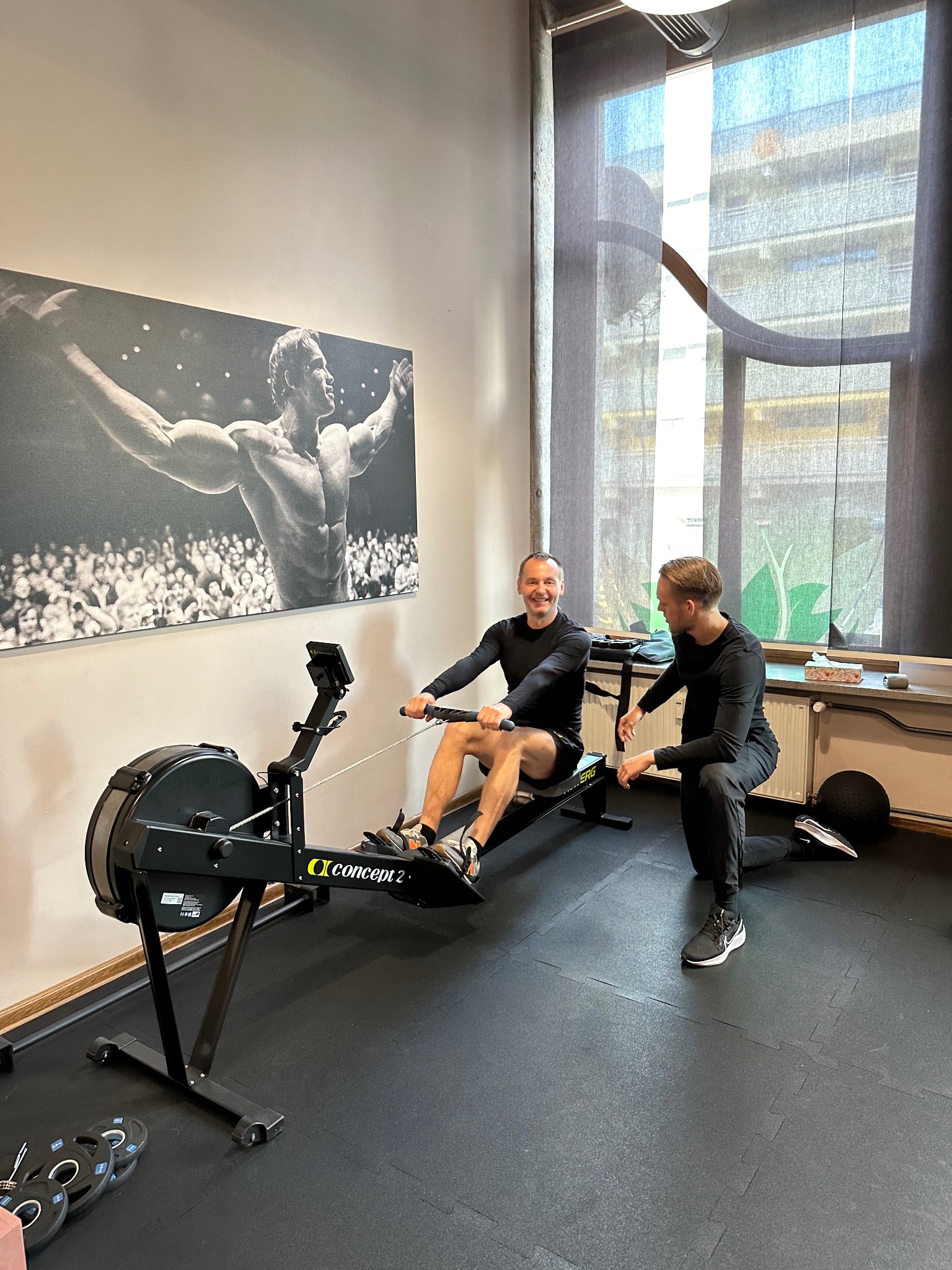 Mężczyzna ćwiczy na ergometrze wioślarskim pod okiem instruktora w siłowni, na ścianie plakat z kulturystą.