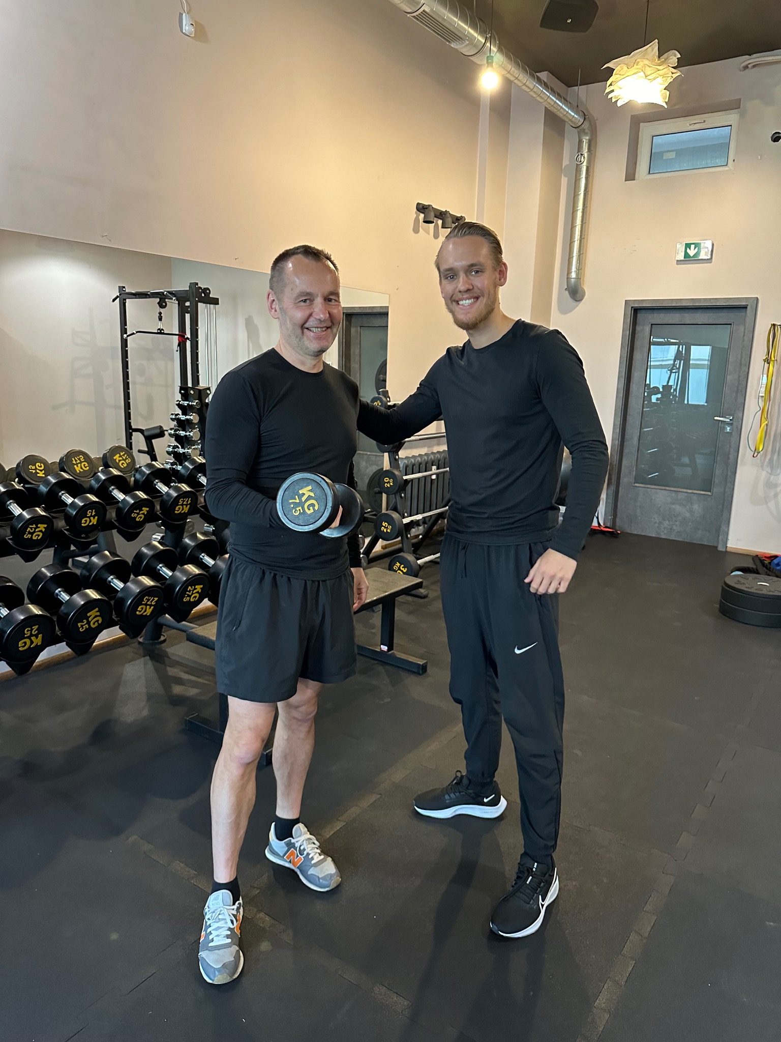Mężczyzna we Wrocławiu ćwiczy z hantlą 7.5 kg pod okiem uśmiechniętego instruktora w siłowni z rzędem hantli na stojaku w tle.