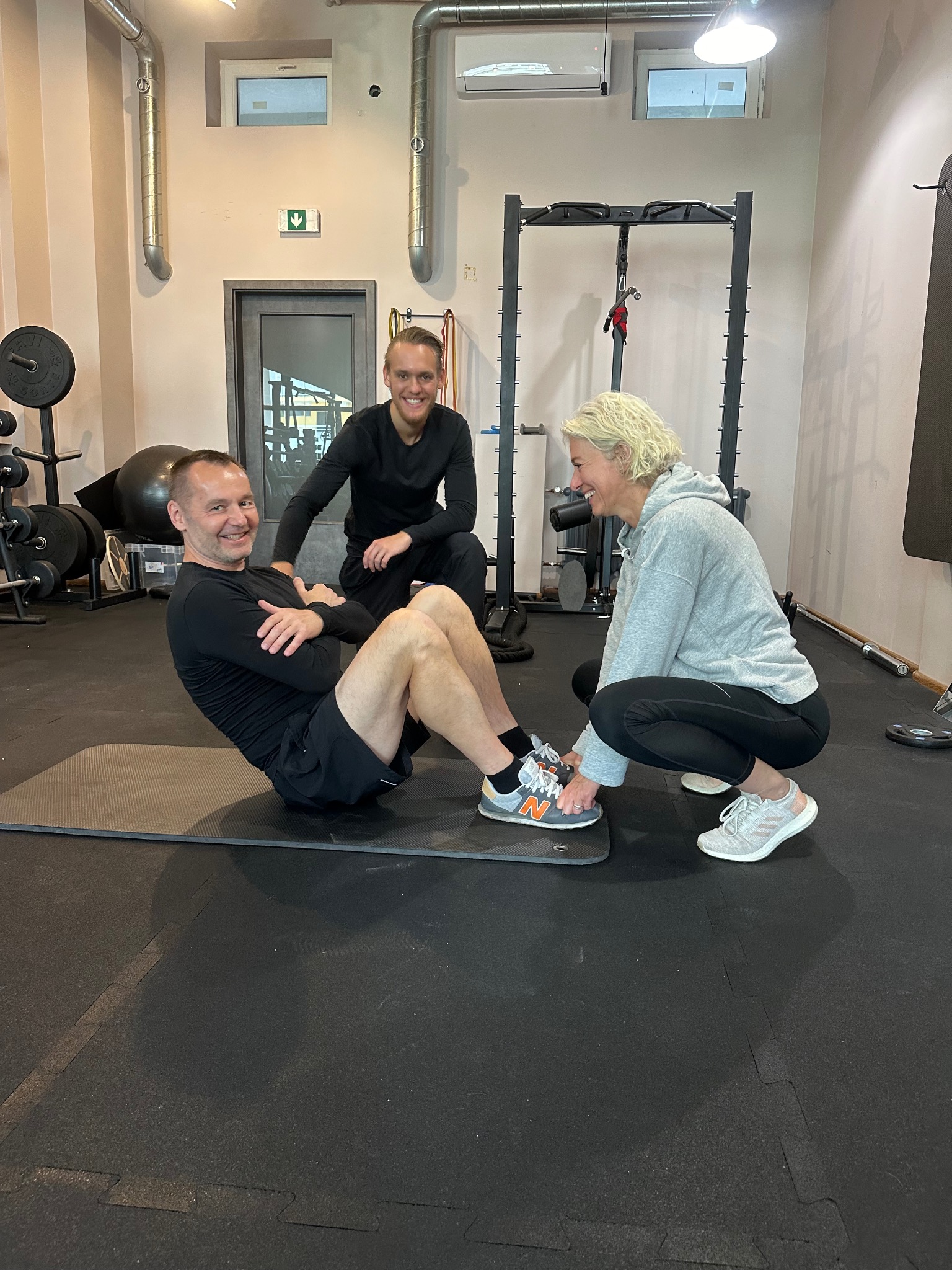 Mężczyzna wykonuje ćwiczenia na brzuchu na macie, kobieta w szarej bluzie trzyma jego stopy, a drugi mężczyzna w czarnej koszulce siedzi za nimi uśmiechnięty, w tle sprzęt do ćwiczeń.