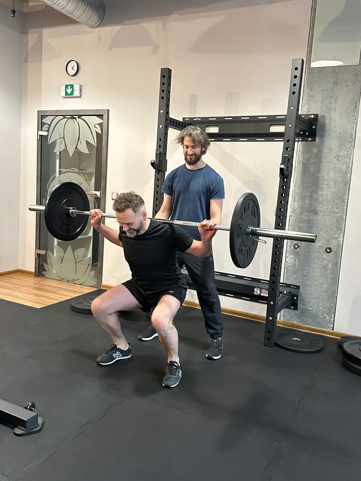 Mężczyzna wykonuje przysiad ze sztangą, a drugi mężczyzna go asekuruje w siłowni z czarną podłogą i sprzętem do ćwiczeń.