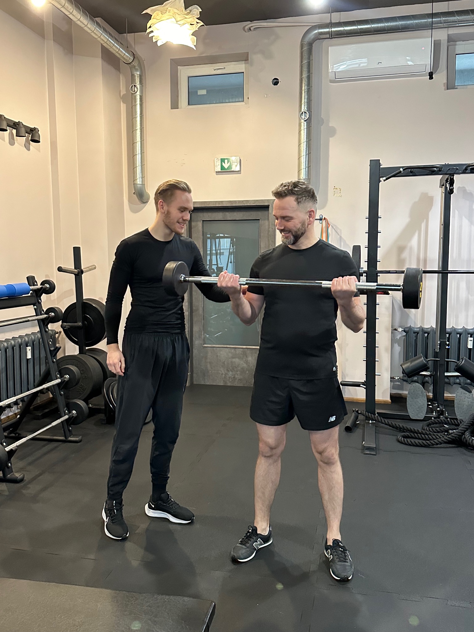 Mężczyzna w czarnym stroju ćwiczy biceps z sztangą pod nadzorem drugiego mężczyzny w siłowni.