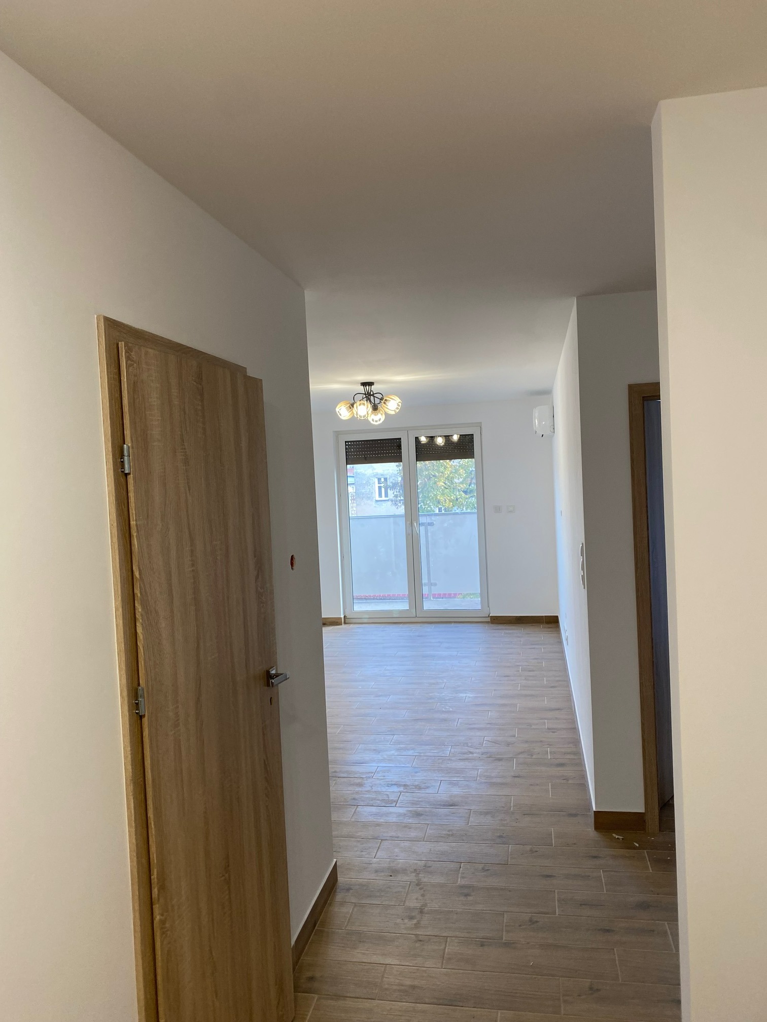 Wykończone wnętrze mieszkania z drewnopodobną podłogą, widok z korytarza na salon z balkonem i nowoczesnym oświetleniem sufitowym, drzwi wewnętrzne z drewnianą okleiną po lewej stronie.