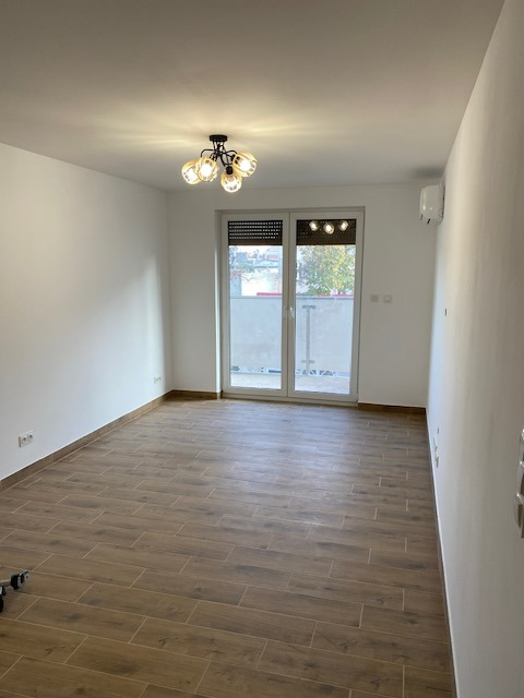 Pusty pokój po remoncie z drewnopodobną podłogą z płytek, białymi ścianami, nowoczesną lampą sufitową i klimatyzacją, widok na balkon przez przeszklone drzwi.