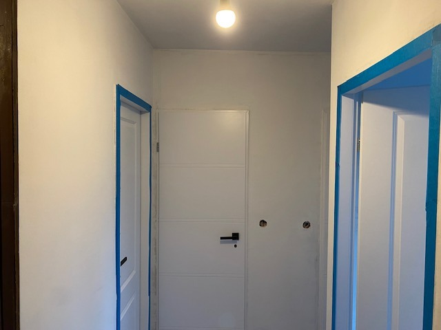 Korytarz po malowaniu na biało, z niebieską taśmą malarską zabezpieczającą ościeżnice drzwi, widoczne ślady po gniazdkach elektrycznych.