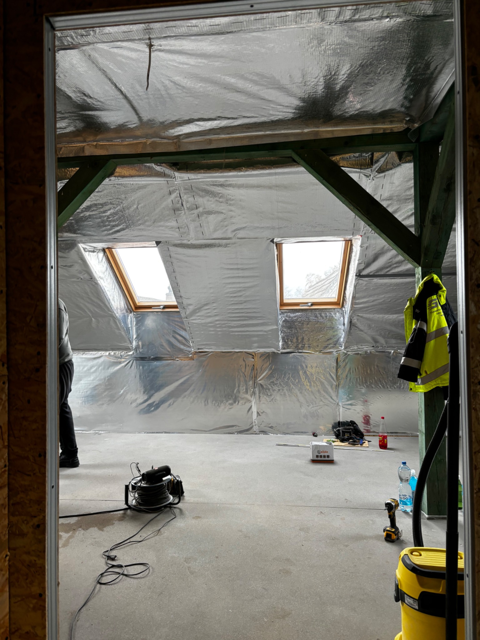 Poddasze w trakcie izolacji folią aluminiową, widoczne okna dachowe, drewniana konstrukcja, narzędzia i odzież robocza rozłożone na podłodze.