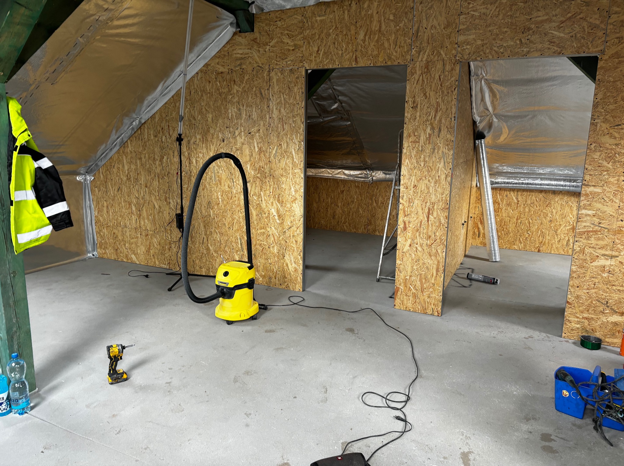 Poddasze w trakcie adaptacji: ściany obite płytami OSB, izolacja folią aluminiową, żółty odkurzacz przemysłowy Kärcher, wiertarka DeWalt na posadzce, widoczne otwory drzwiowe i drabina w tle.