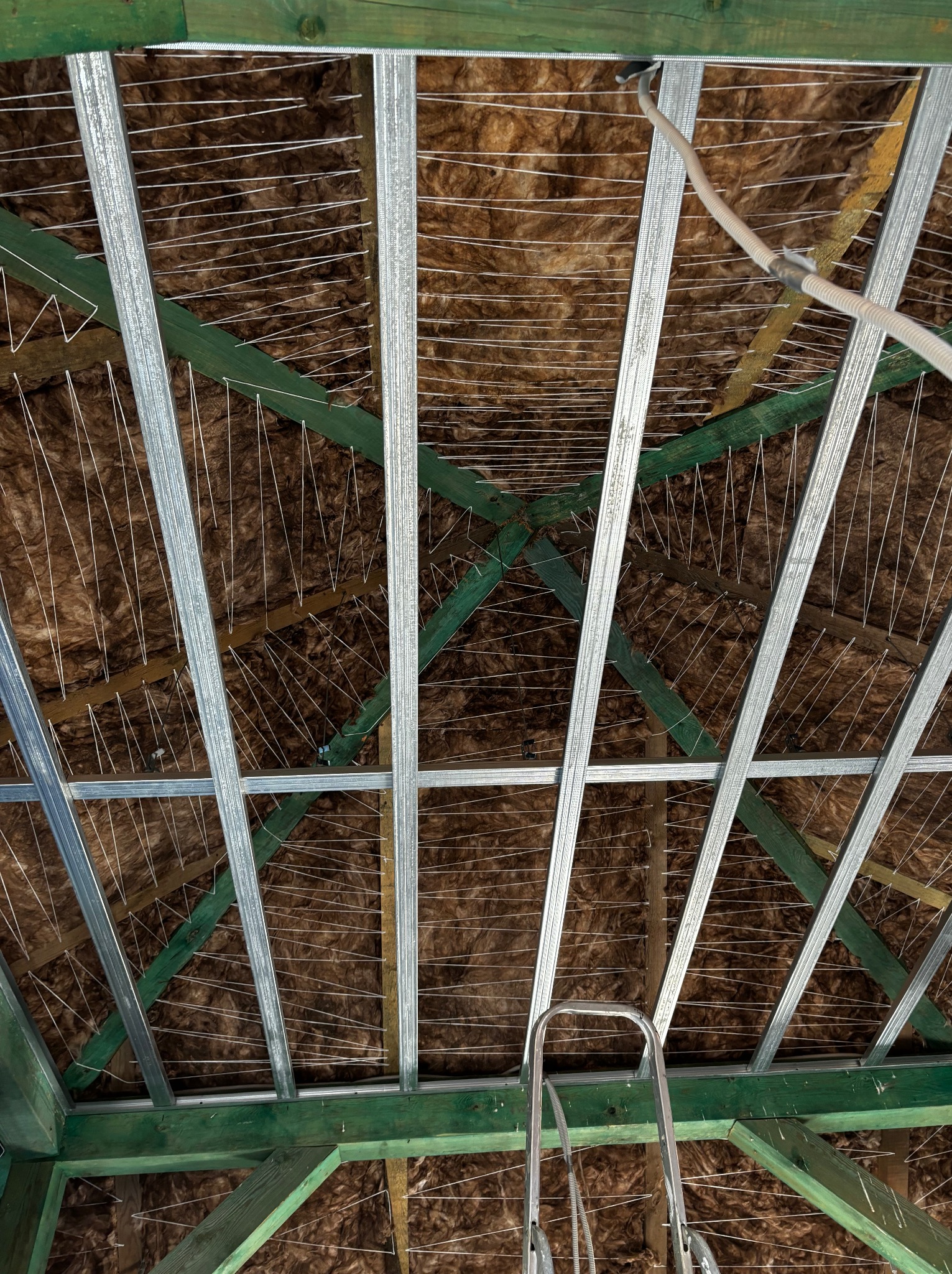 Montaż izolacji z wełny mineralnej w poddaszu, widoczne metalowe profile pod płyty karton-gips, drewniana konstrukcja dachu pomalowana na zielono i częściowo widoczna drabina.