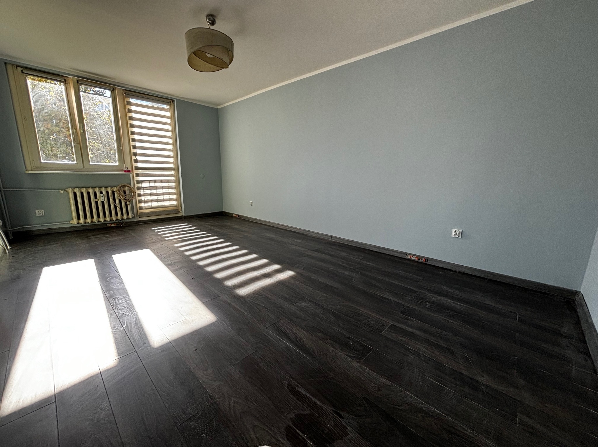 Pomalowane na szaro mieszkanie z ciemną podłogą, grzejnikiem pod oknem z roletami i lampą sufitową.