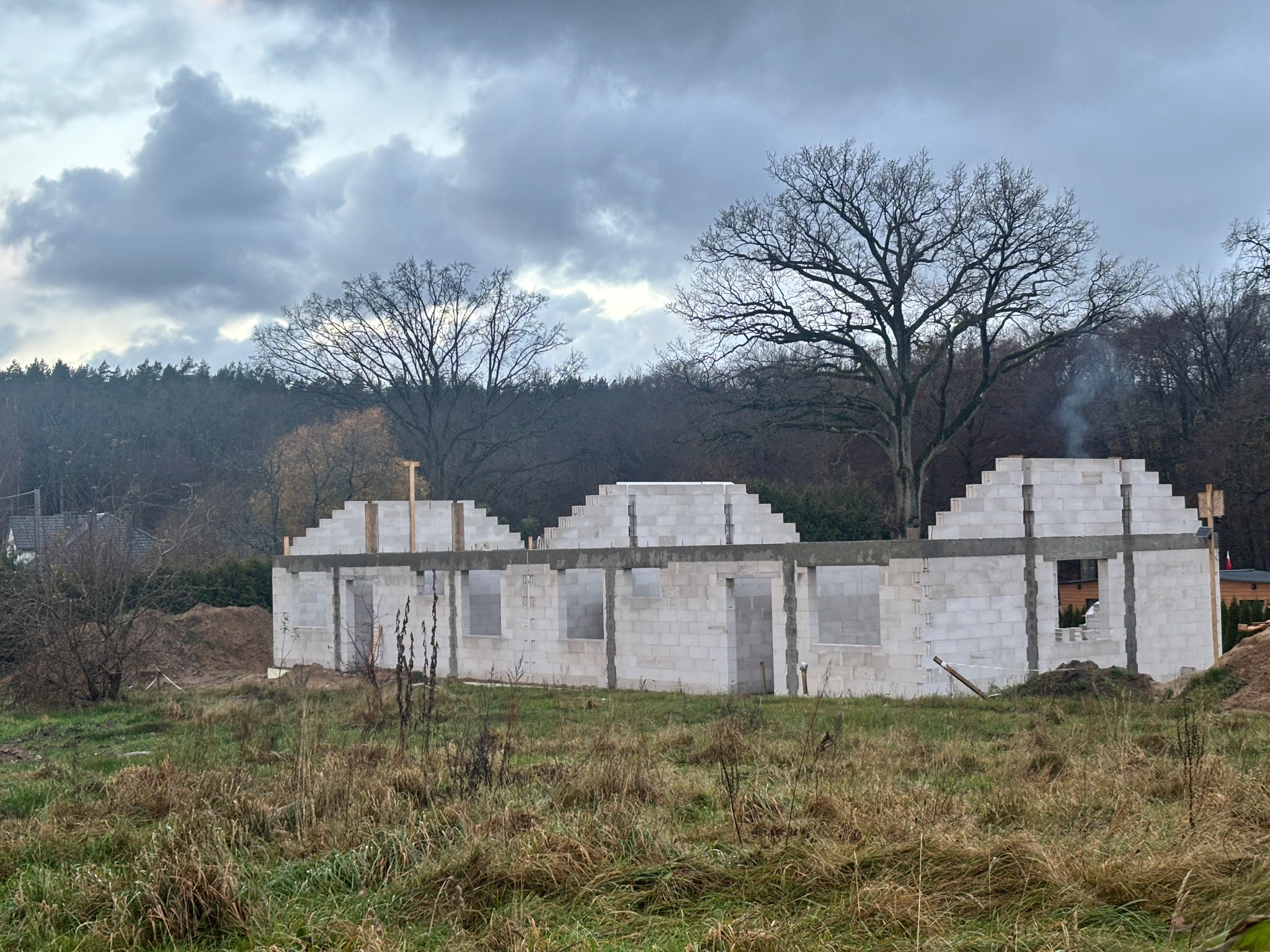 Wznoszenie ścian domu z bloczków betonu komórkowego na tle pochmurnego nieba i drzew. Widoczne otwory okienne i drzwiowe, teren budowy z trawą.