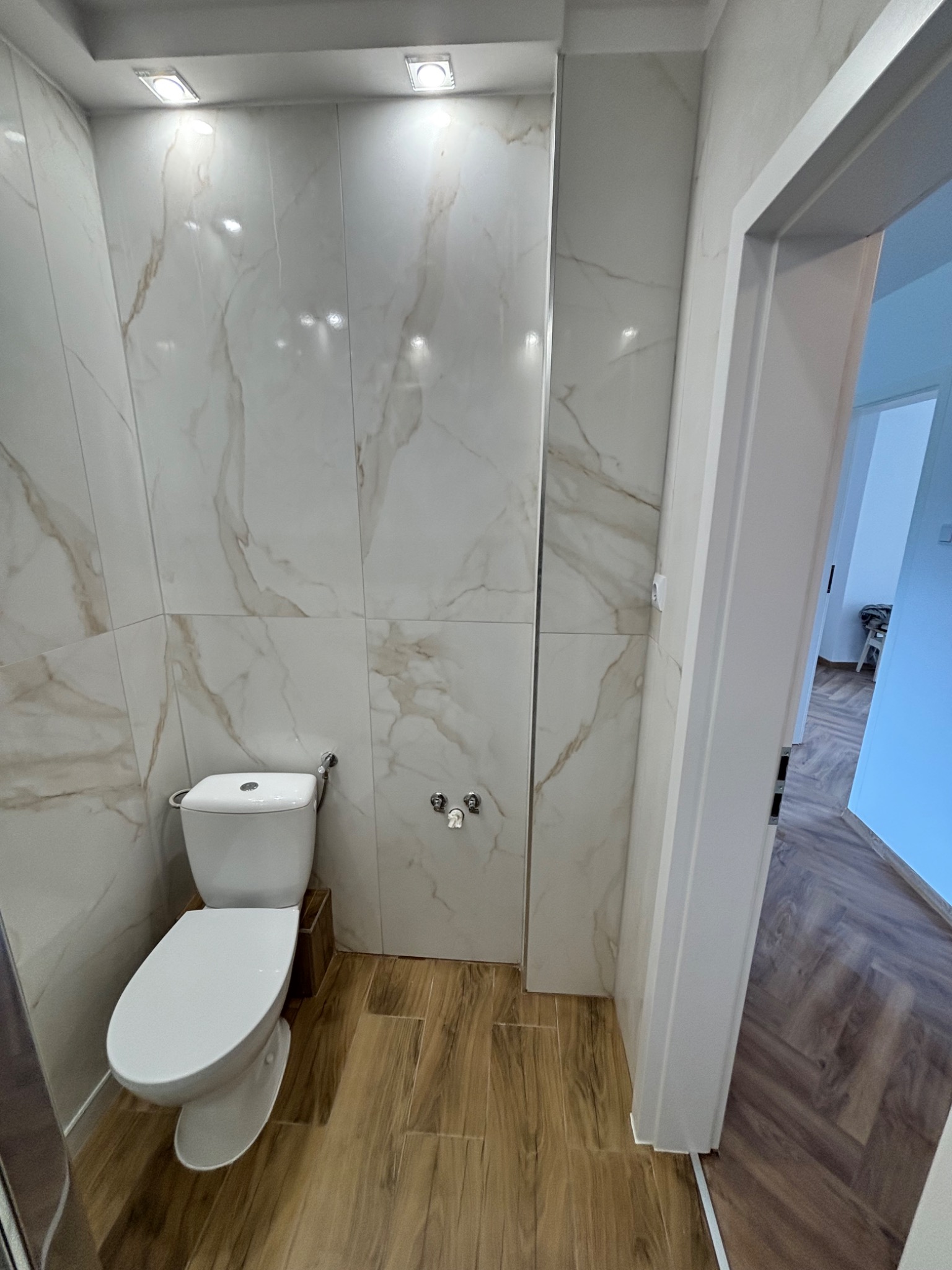 Wykończona łazienka z toaletą, płytkami imitującymi marmur na ścianach i drewnopodobną podłogą. Widoczne oświetlenie sufitowe i fragment otwartego przejścia do innego pomieszczenia.
