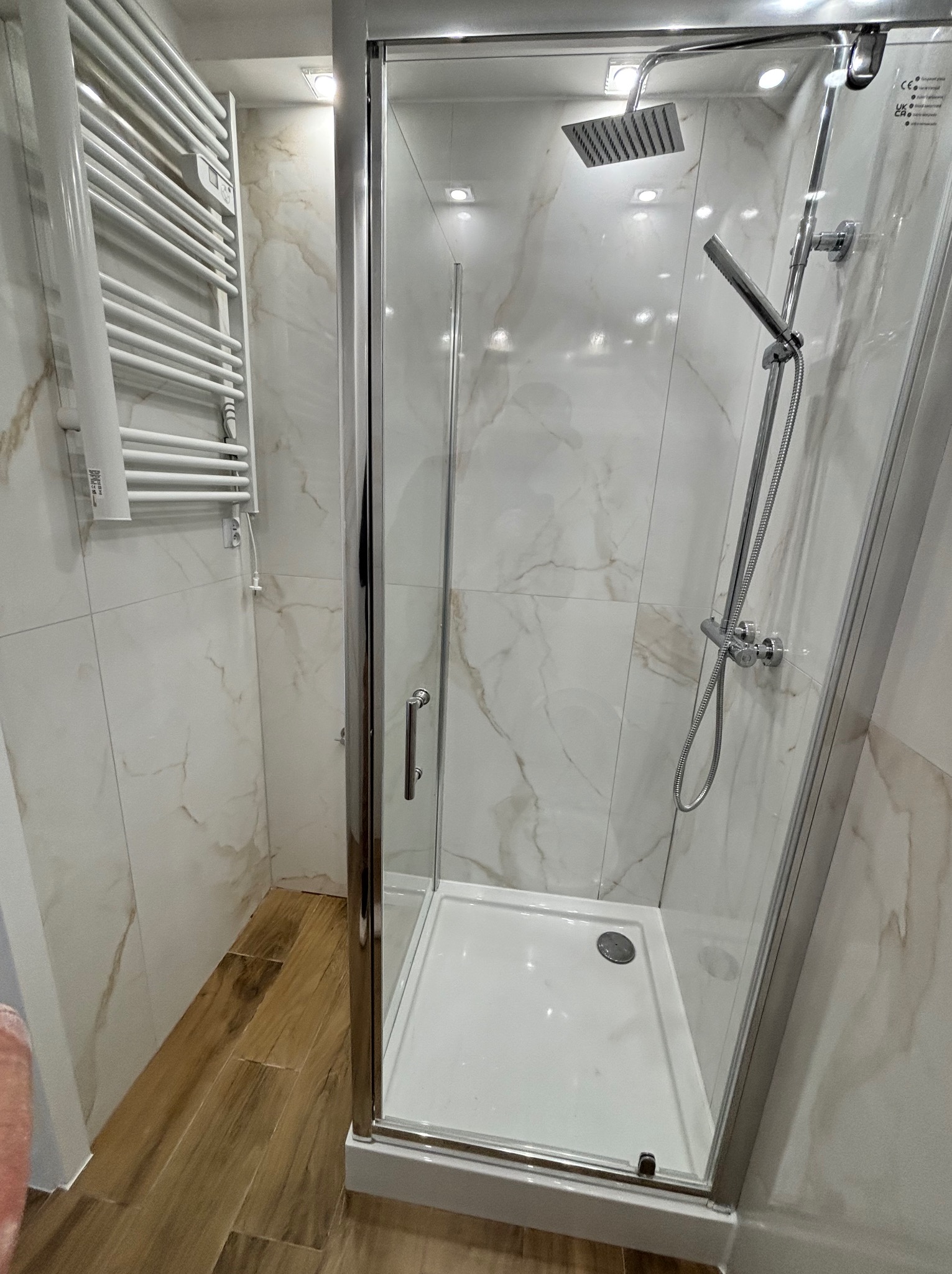 Nowoczesna kabina prysznicowa z deszczownicą i grzejnikiem drabinkowym na tle marmurowych płytek. Jasne wnętrze łazienki po remoncie, widok z boku.