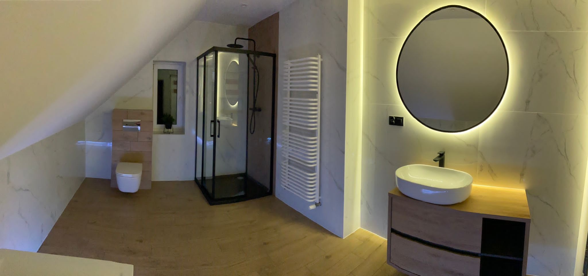 Nowoczesna łazienka na poddaszu z kabiną prysznicową w czarnej ramie, okrągłym lustrem z podświetleniem LED, umywalką nablatową na drewnianej szafce i grzejnikiem drabinkowym.