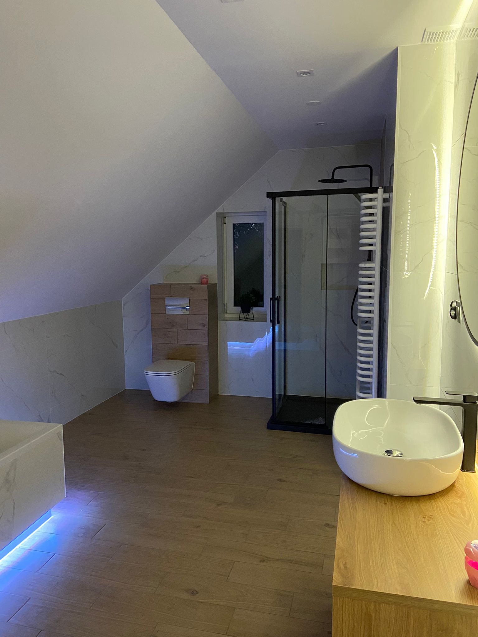 Nowoczesna łazienka na poddaszu z kabiną prysznicową typu walk-in, umywalką nablatową na drewnianym blacie, podświetlaną wanną oraz toaletą w zabudowie z drewnianymi panelami.