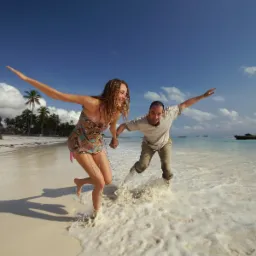 Para trzymająca się za ręce, biegnąca po piaszczystej plaży w tropikach, rozbryzgująca wodę, z tradycyjną łodzią na horyzoncie i palmami w tle.