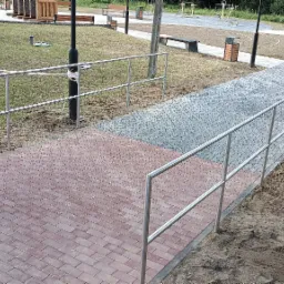 Nowo wybudowany chodnik z kostki brukowej w dwóch odcieniach szarości i czerwieni z zainstalowanymi metalowymi barierkami ochronnymi na obrzeżach, część terenu jeszcze niezagospodarowana.