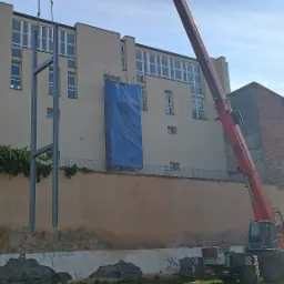 Montaż stalowej konstrukcji przy użyciu dźwigu na tle budynku z odsłoniętą cegłą i pnącą się roślinnością.