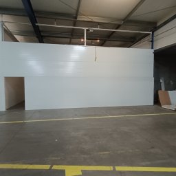 Polconstruction - Wnętrze hali produkcyjnej z zamontowanymi białymi płytami warstwowymi i wyciętym otworem drzwiowym, widoczne żółte linie na podłodze, w tle składowane płyty i fragment sylwetki mężczyzny.